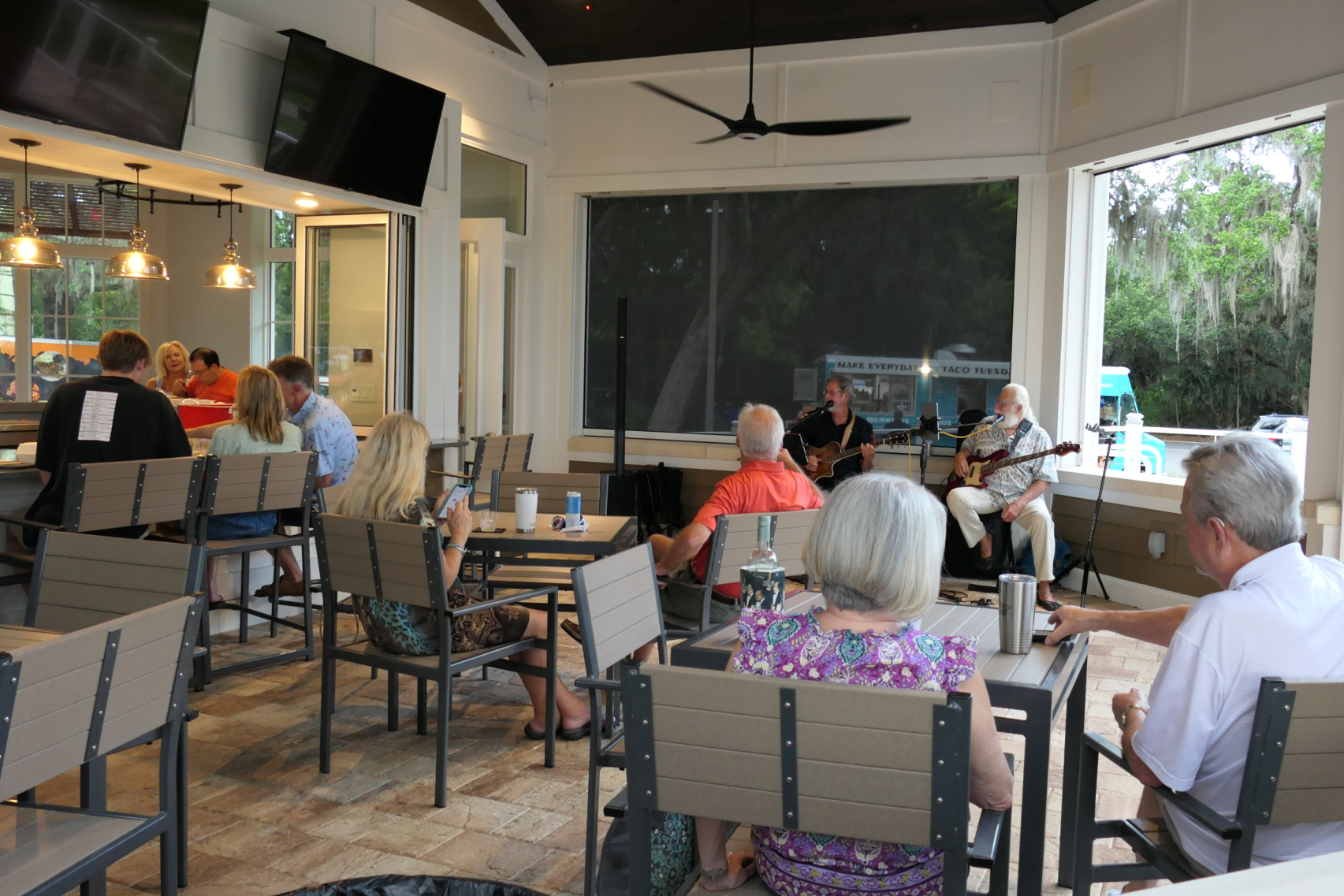 People sitting on the screened porch at The Oaks listening to a duo performing. A food truck is visible behind the performers.